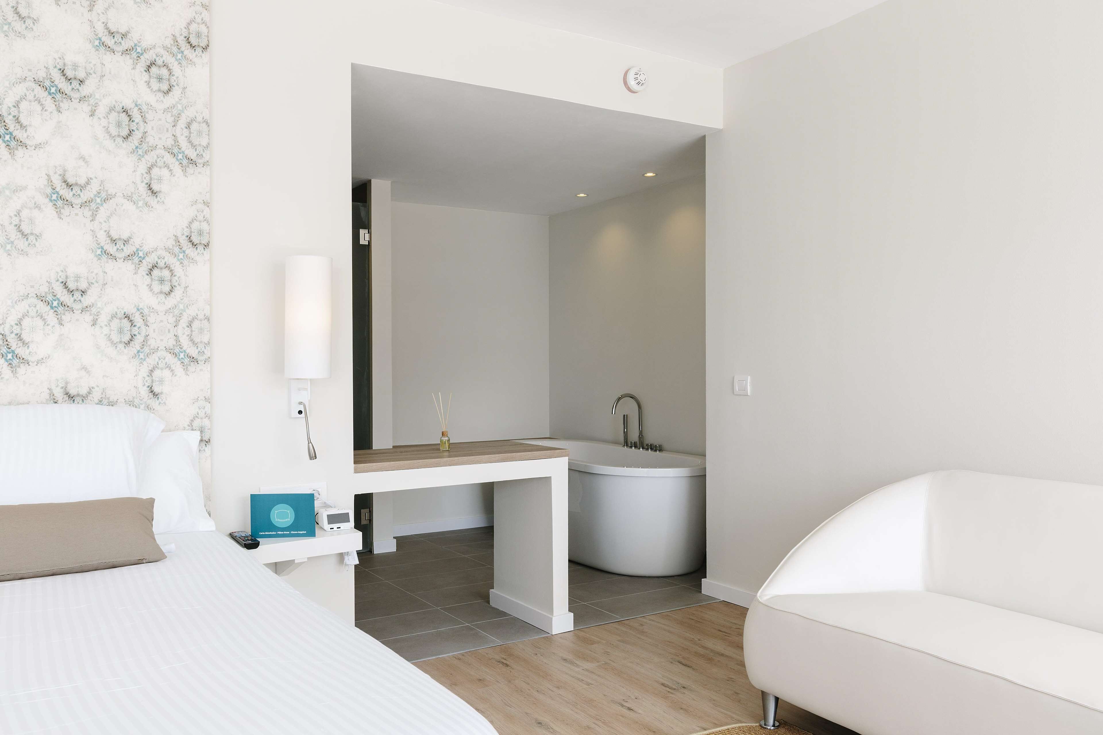 A modern hotel room with a bed, a white sofa, a desk with a sink, and a bathtub.