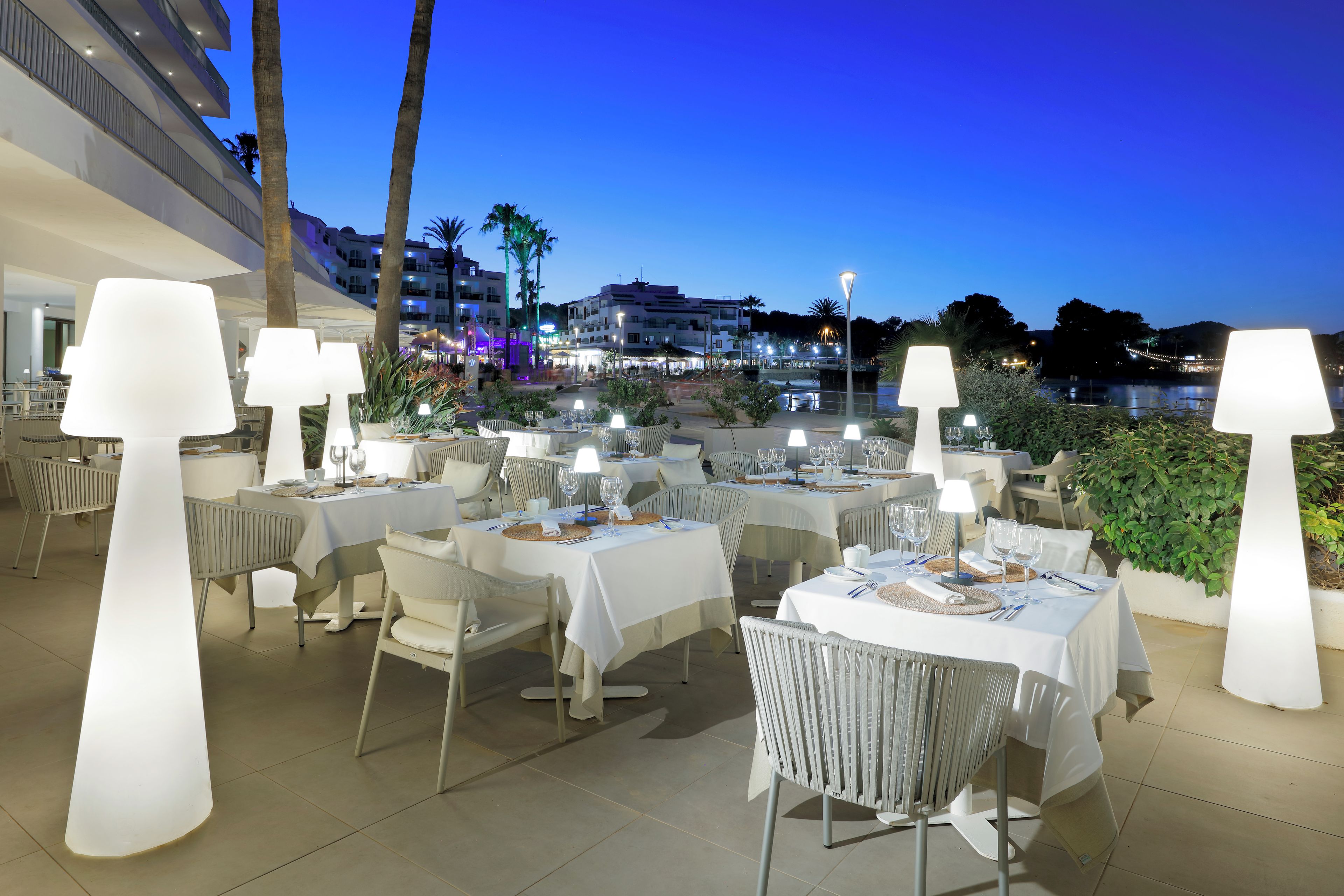 Un área de comedor al aire libre con mesas dispuestas para dos, cada una con mantel blanco y lámpara.