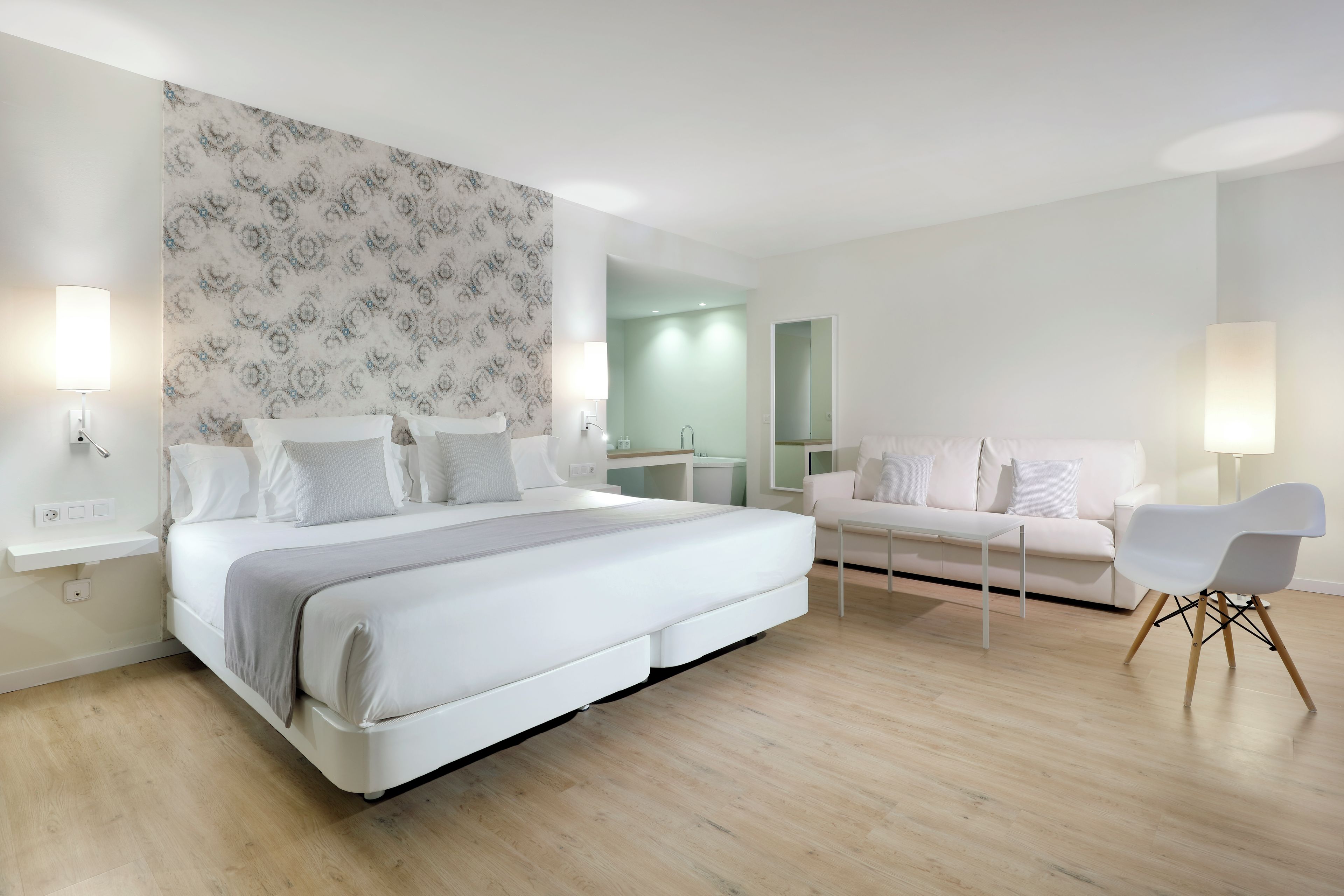 Una habitación de hotel moderna con una cama grande, un rincón de descanso con un sofá y una silla, una mesita y un baño visible a través de una puerta abierta.