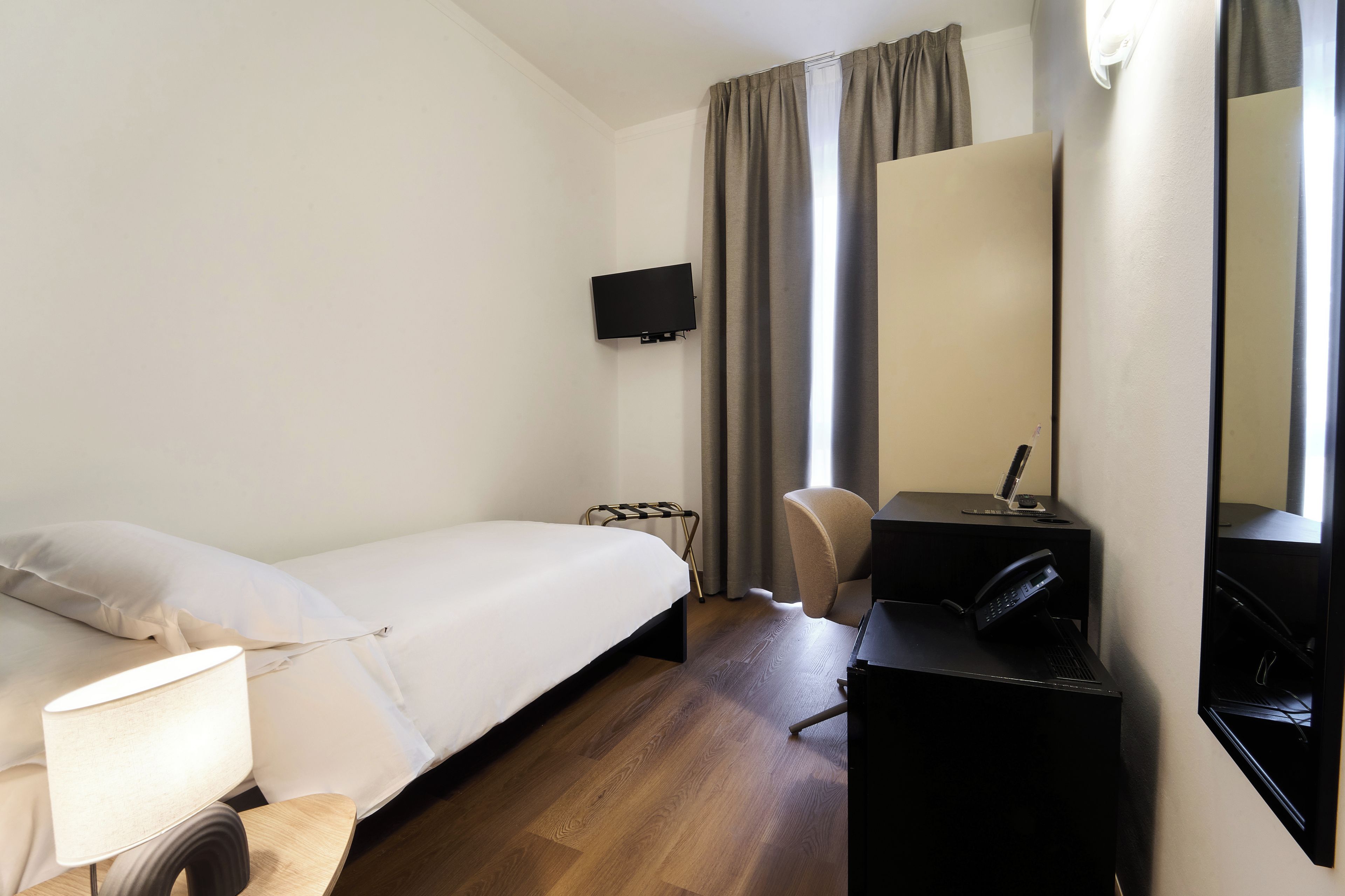 Une chambre d’hôtel comprenant un lit, un bureau, une chaise, une lampe de bureau, un téléphone, une télévision et une fenêtre avec des rideaux.