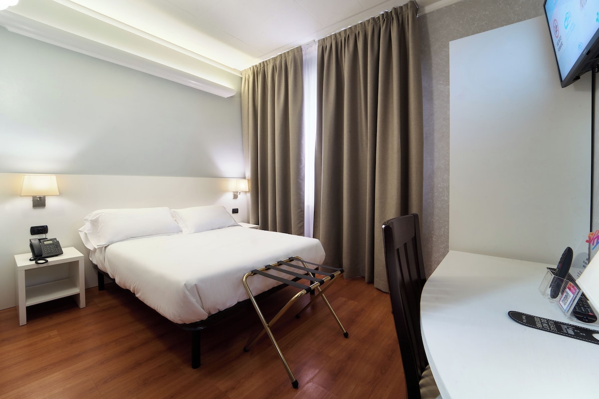 Een hotelkamer met een bed, nachtlampjes, een bureau met een stoel, een televisie en een raam met gordijnen.
