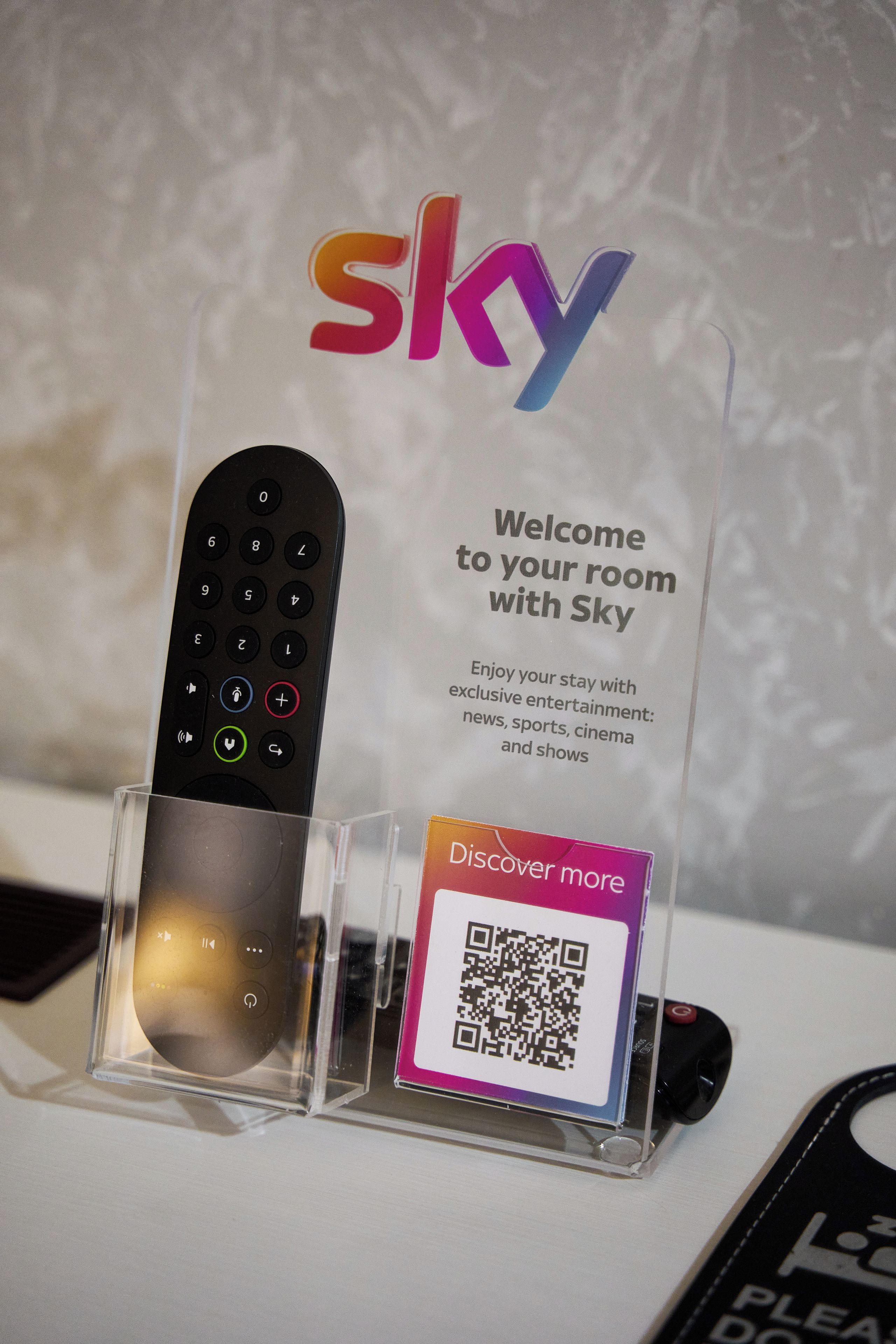 A remote control in a clear stand with a sign above it that reads 'Welcome to your room with Sky' and a QR code below.