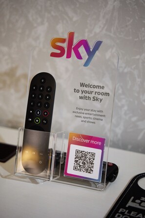 Eine Fernbedienung in einer transparenten Halterung, über ihr ein Schild mit der Aufschrift 'Willkommen in Ihrem Zimmer mit Sky' und darunter ein QR-Code.