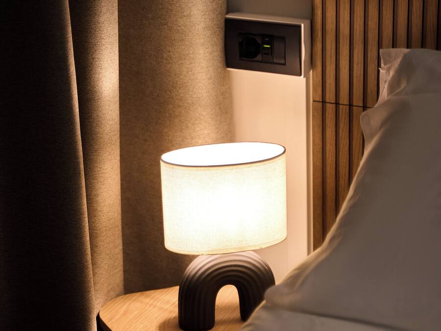 Een nachtkastje met een lamp, een bedieningspaneel aan de muur en een bed met wit beddengoed.