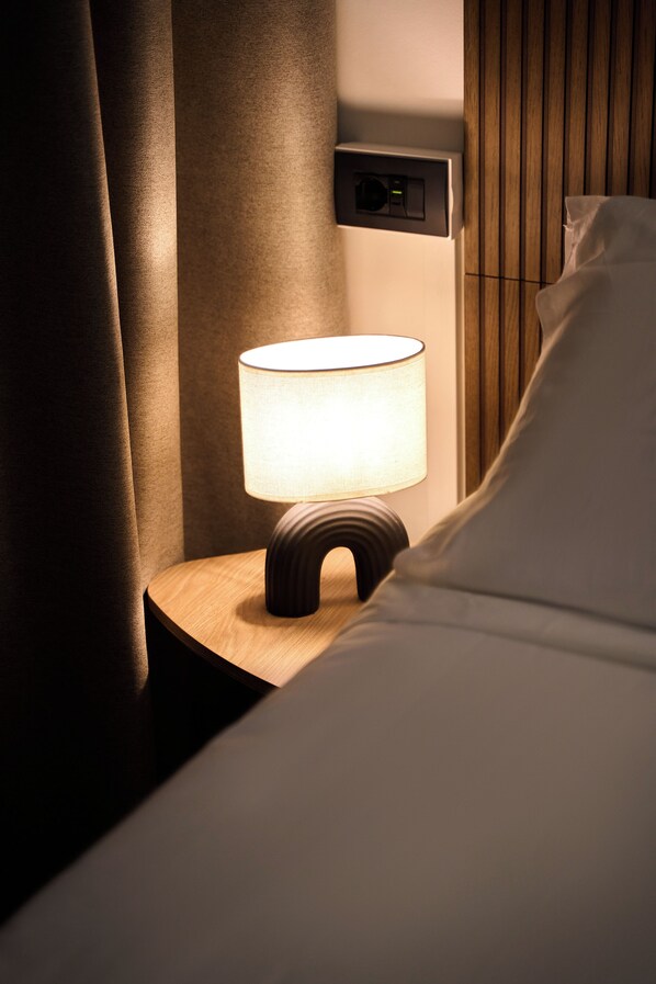 A bedside table with a lamp, a wall-mounted control panel, and a bed with white bedding.