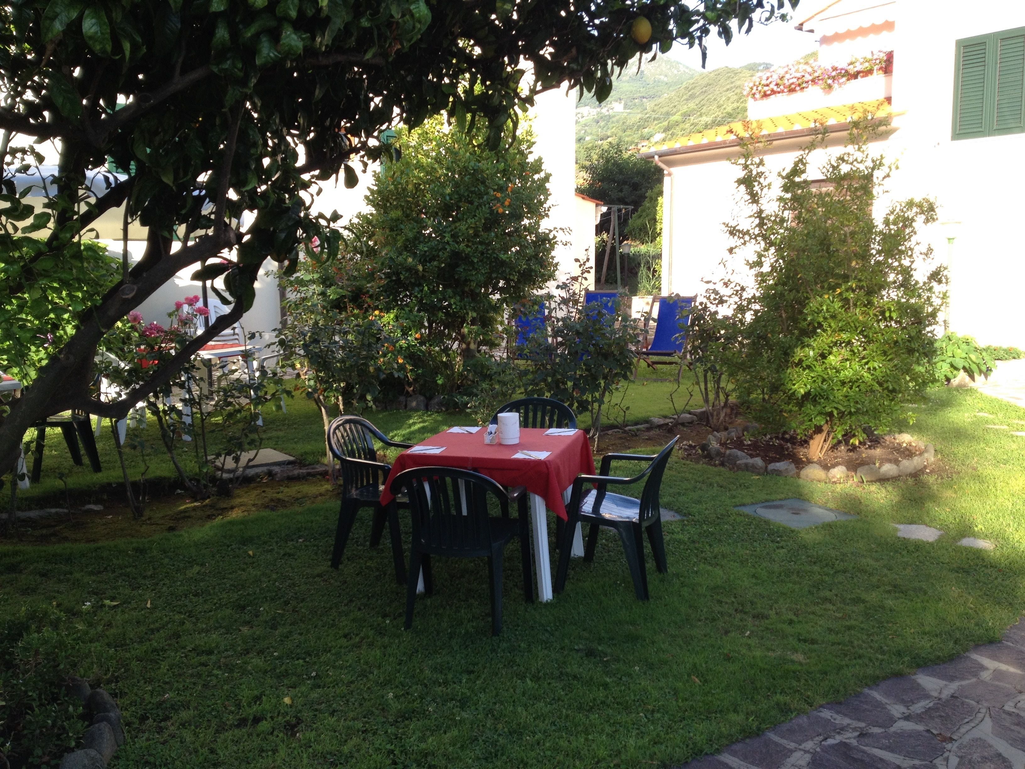 Un espace extérieur aménagé pour les repas, avec une nappe rouge, des chaises noires et un chemin de pierre menant à un bâtiment à la façade blanche et aux volets verts.