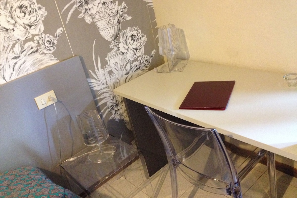 Una habitación con una mesa blanca, sillas transparentes, un papel tapiz floral y una carpeta roja sobre la mesa.