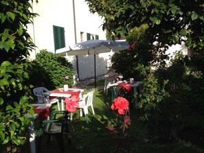 Une terrasse extérieure avec des tables et des chaises, entourée de verdure et avec un bâtiment blanc en arrière-plan.