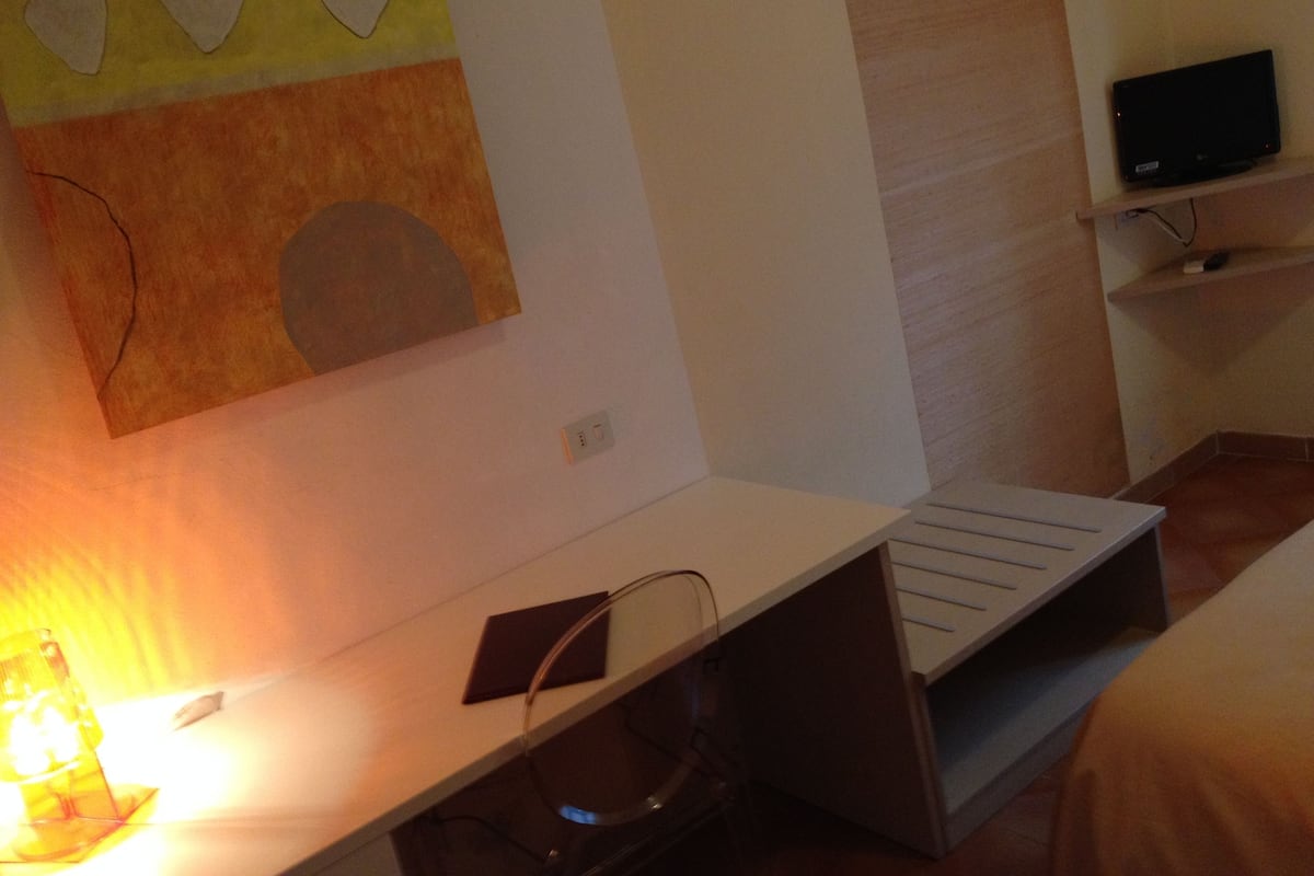 Habitación de hotel con un escritorio blanco, un televisor de pantalla plana sobre una estantería y una pared con un cuadro geométrico.
