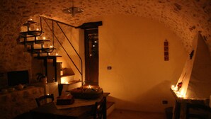 Une pièce faiblement éclairée, avec un mur en pierre rustique, une table en bois, des chaises, un petit escalier et une seule lampe suspendue.
