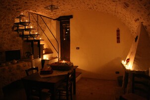 Una habitación tenuemente iluminada con una pared de piedra rústica, una mesa de madera, sillas, una pequeña escalera y una única lámpara colgante.