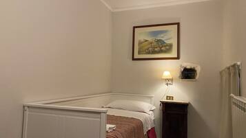 Ein kleines Zimmer mit einem Einzelbett, einem weißen Kopfteil, einer Nachttischlampe, einem gerahmten Bild an der Wand und einem kleinen Schrank.