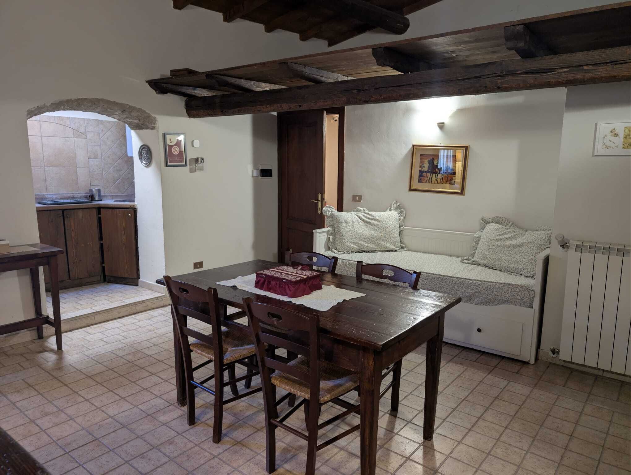 Un coin repas accueillant avec une table et des chaises en bois, un canapé recouvert d’une couverture à motifs floraux et une cuisine visible à travers une arche.