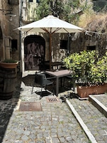 Un patio exterior con suelo de piedra, una mesa y sillas, un gran paraguas y plantas en macetas.