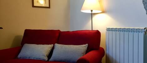 Un sofá rojo con dos almohadones, una lámpara de pie, un cuadro enmarcado y un radiador.