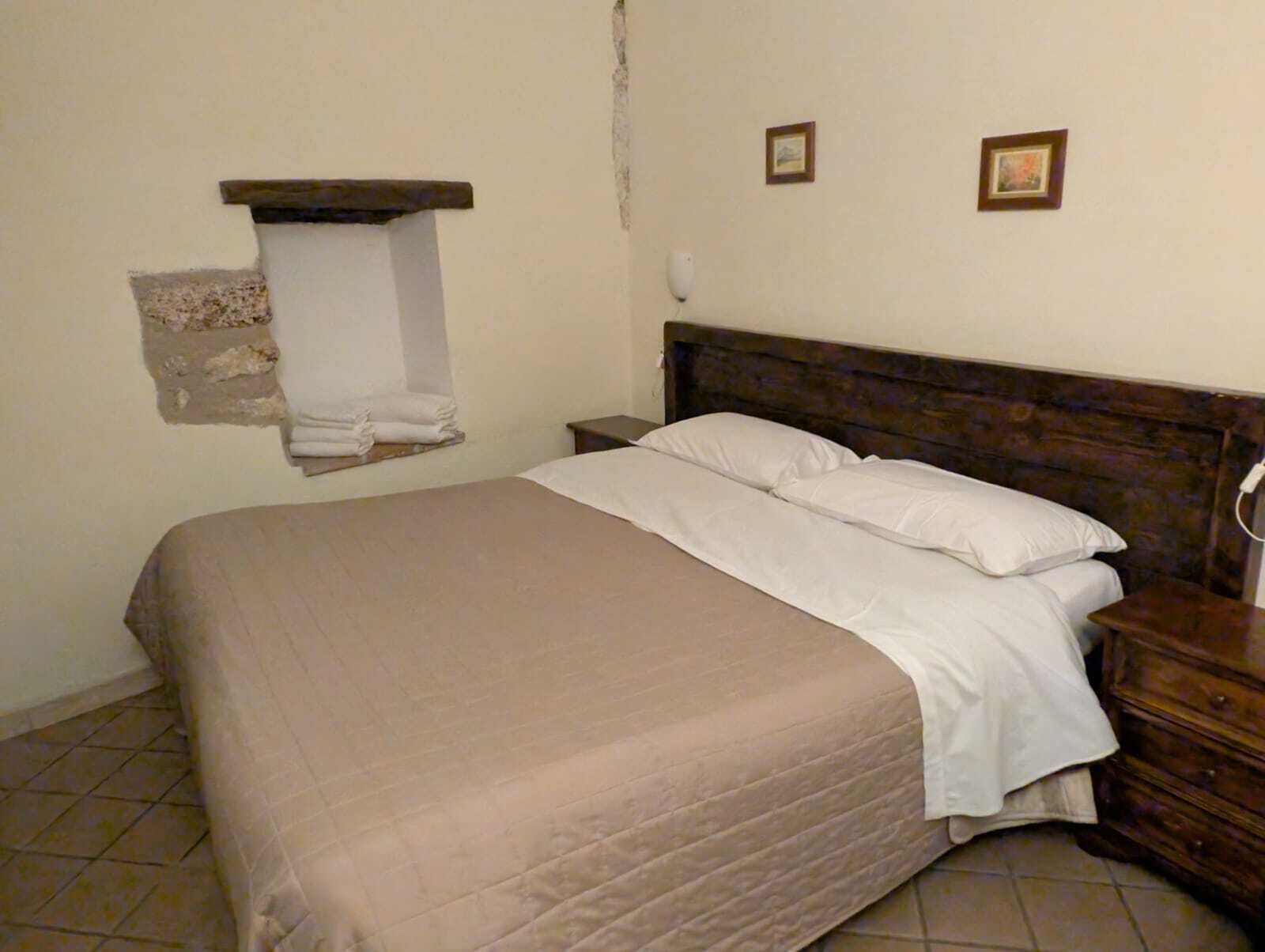 Ein Einzelbett mit Holz-Kopfstütze in einem Zimmer mit schlichten Wänden und gefliestem Boden.