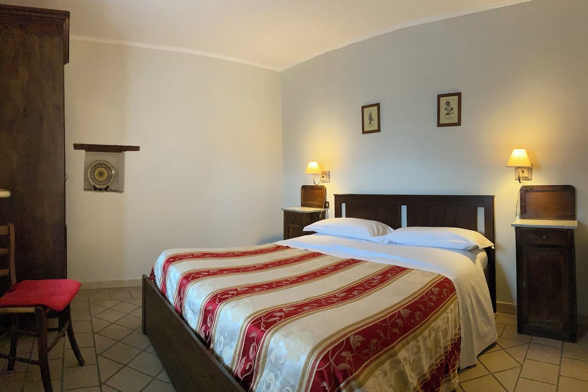 Ein Hotelzimmer mit einem Bett, zwei Nachttischlampen, einer kleinen Ablage mit einer dekorativen Platte und einem Sessel mit rotem Kissen.