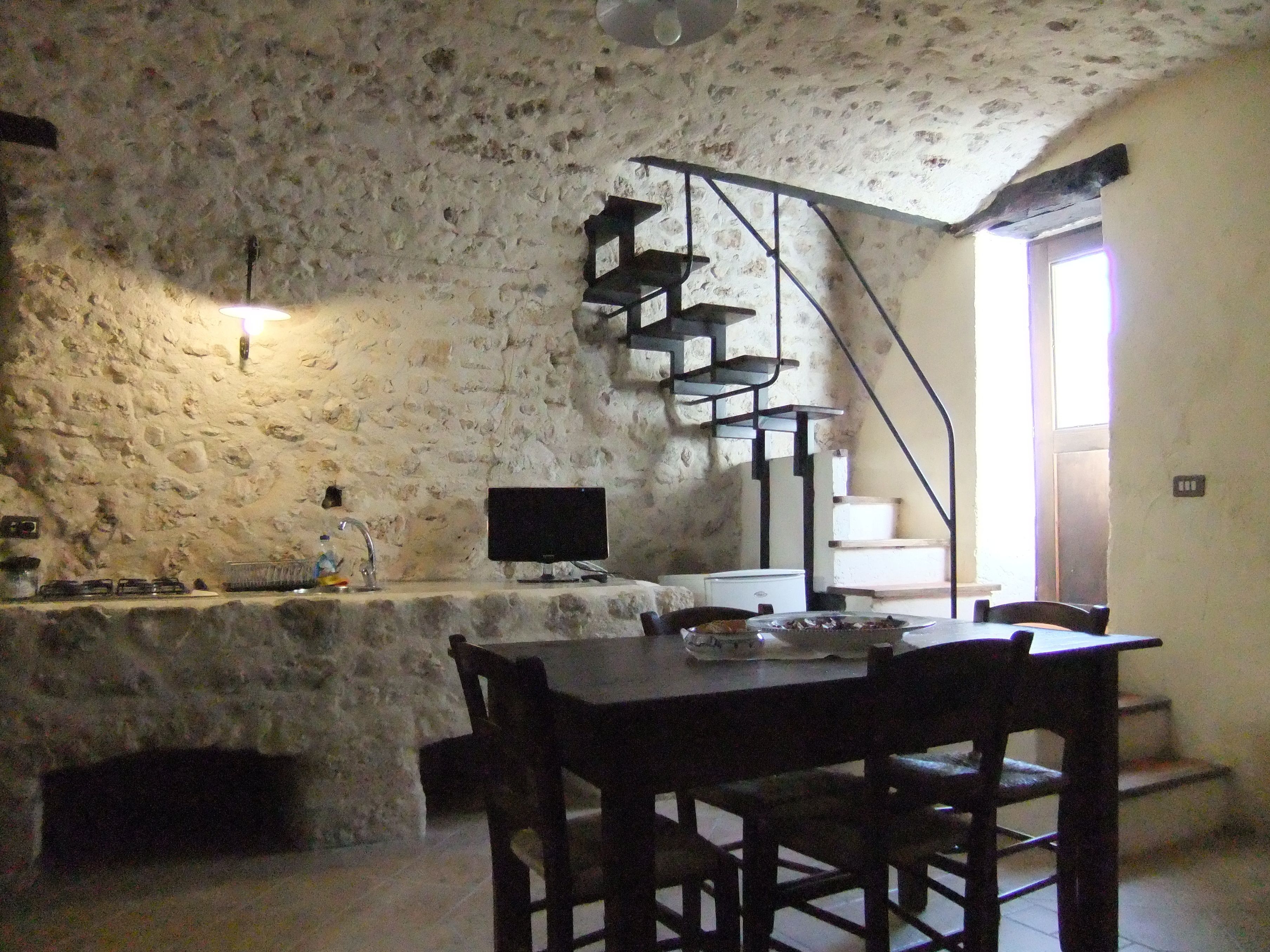 Una cocina rústica con pared de piedra, una mesa de comedor de madera y una escalera que sube a un nivel superior.
