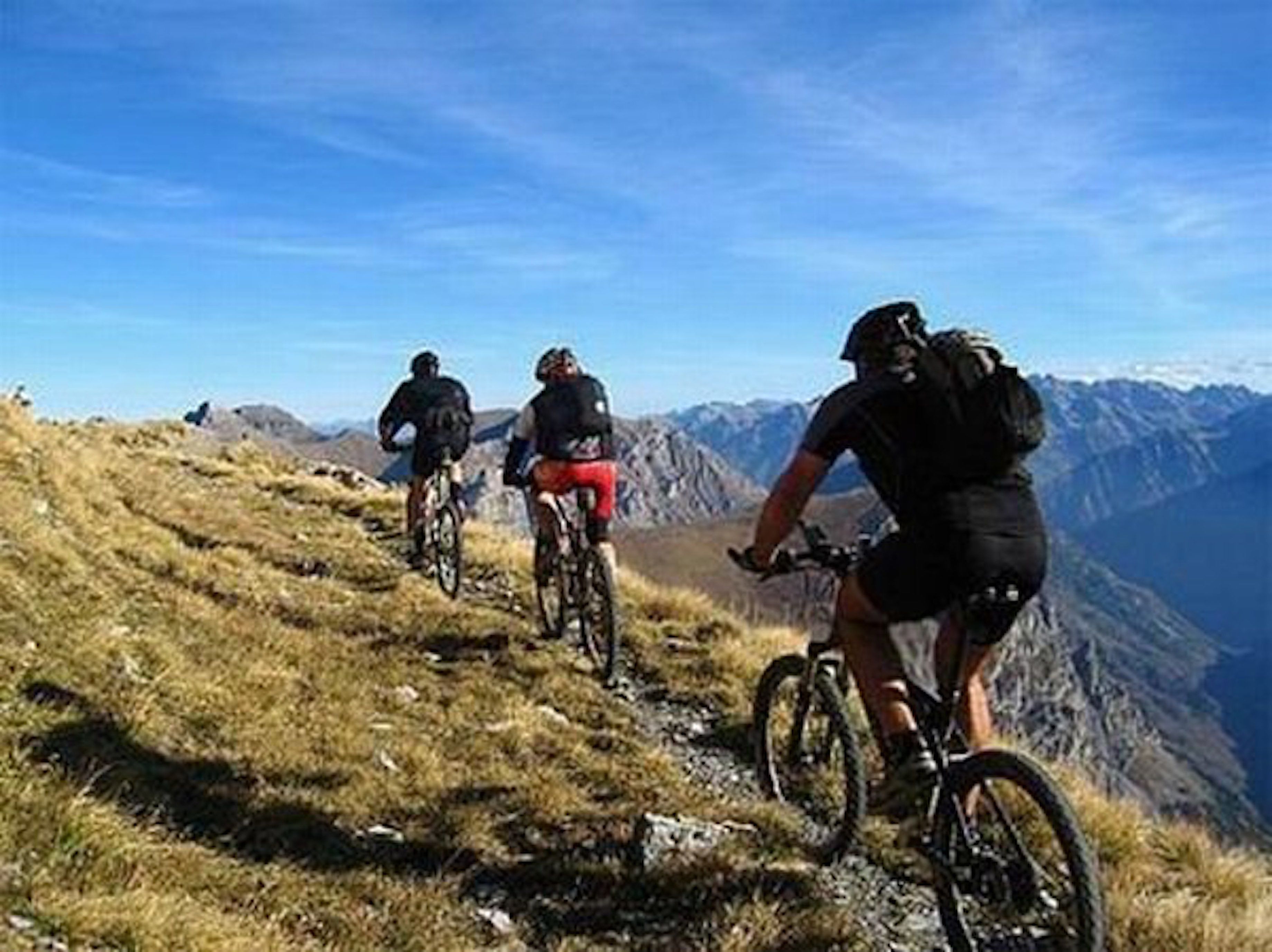 Três ciclistas numa trilha de montanha, com vista clara das montanhas ao redor.