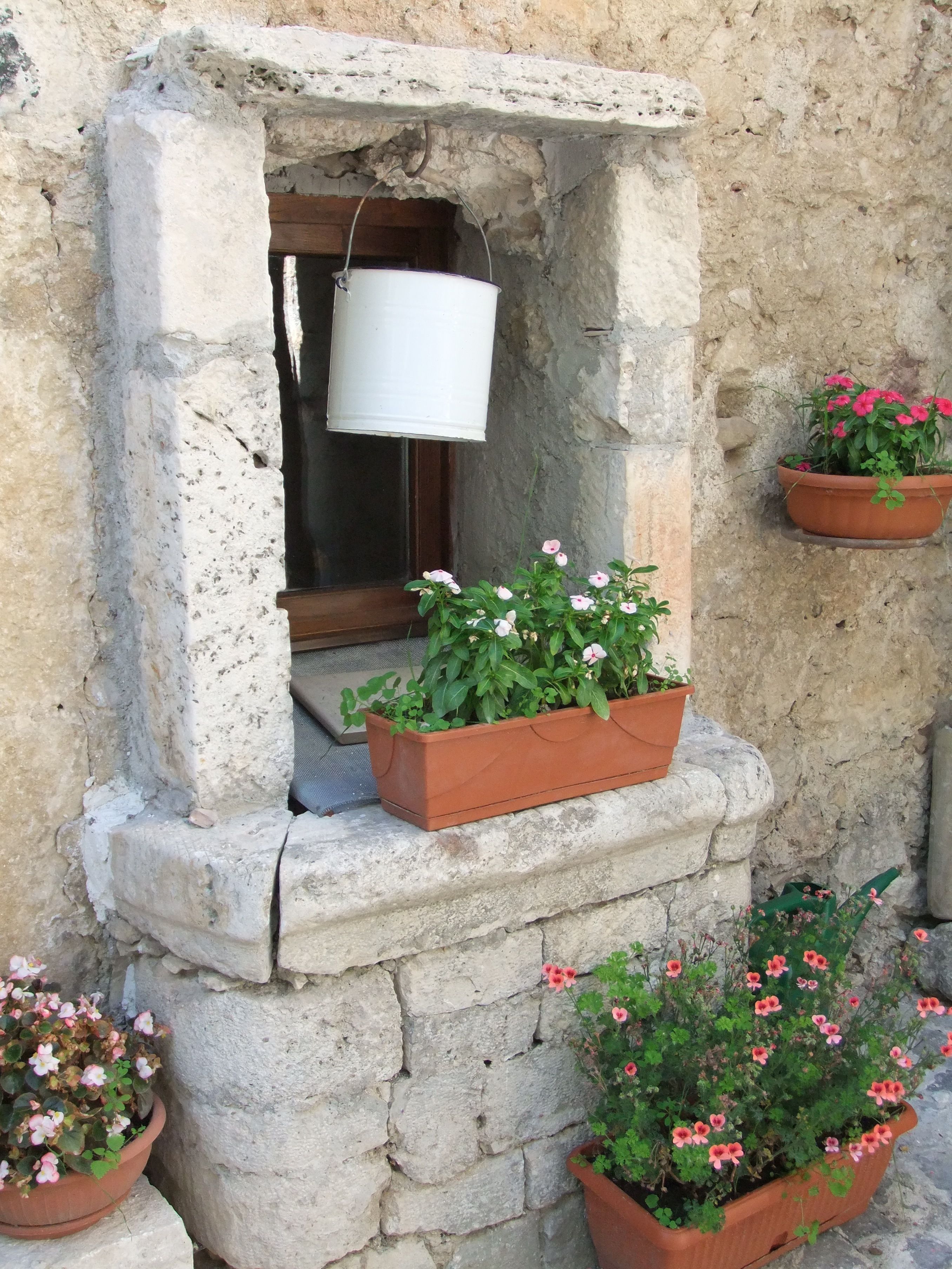 Una ventana de piedra con un balde blanco colgando en su interior, enmarcada por macetas con flores rosadas.