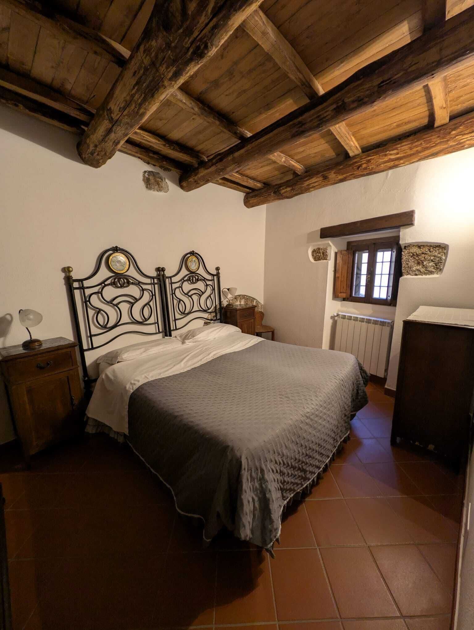 Ein Schlafzimmer mit Holzbalkendecke, einem Bett mit Metallkopfteil, einem Nachttisch, einer Kommode und einem Fenster mit Ausblick.