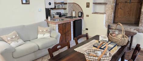 A cozy living room with a sofa, a dining table with chairs, a kitchen area with a sink and stove, and a rustic stone wall.