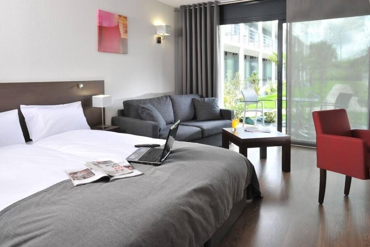 Een moderne hotelkamer met een groot bed, een grijze bank, een rode fauteuil en een laptop op het bed.