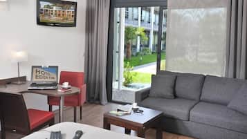 A hotel room with a grey sofa, a small table with a coffee cup, a laptop, and a phone on a bed.