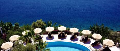 A poolside area with lounge chairs and umbrellas, surrounded by greenery and overlooking the sea.