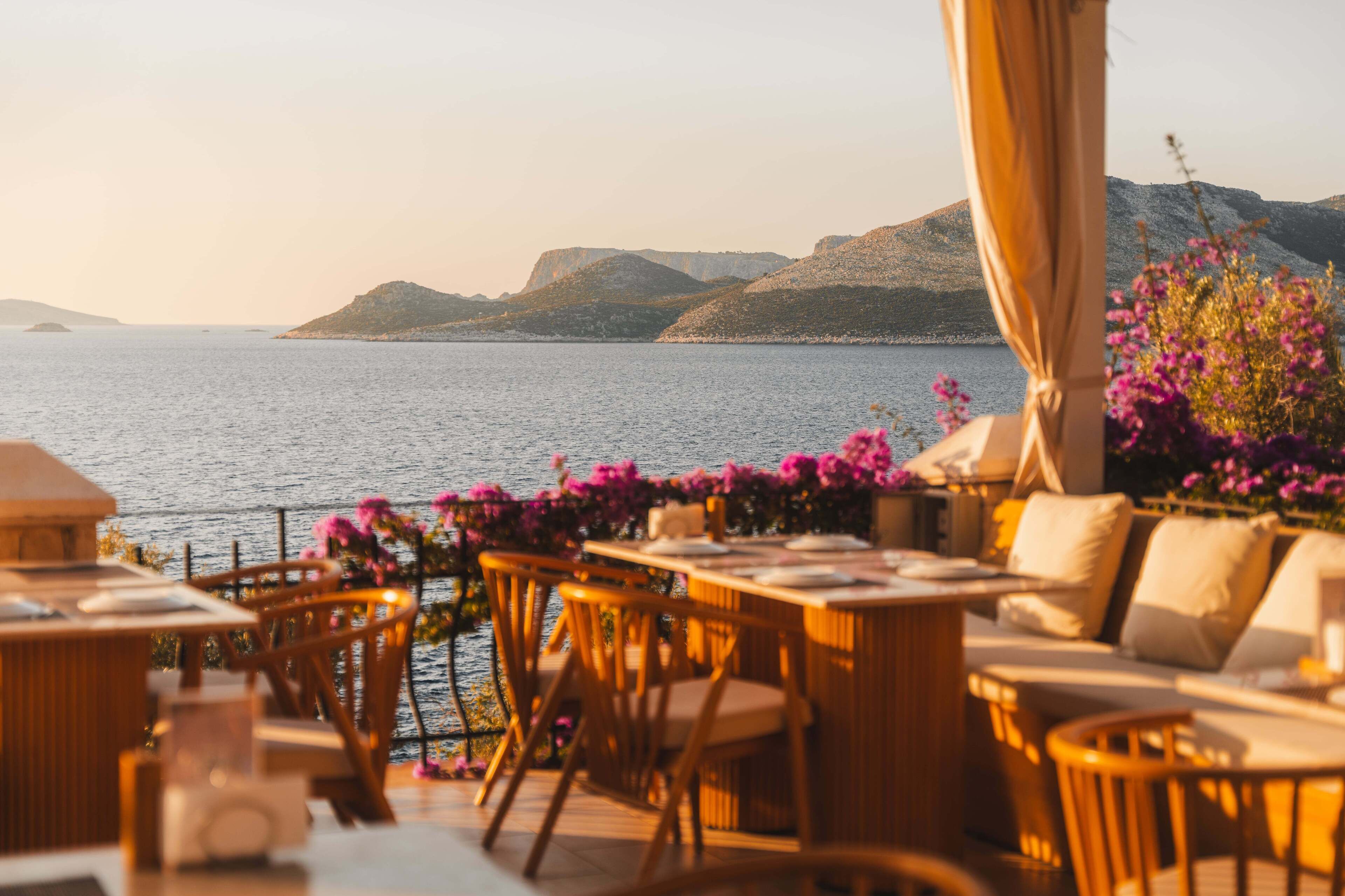 Un espacio de comedor al aire libre con mobiliario de mimbre y vistas al mar y a las colinas.