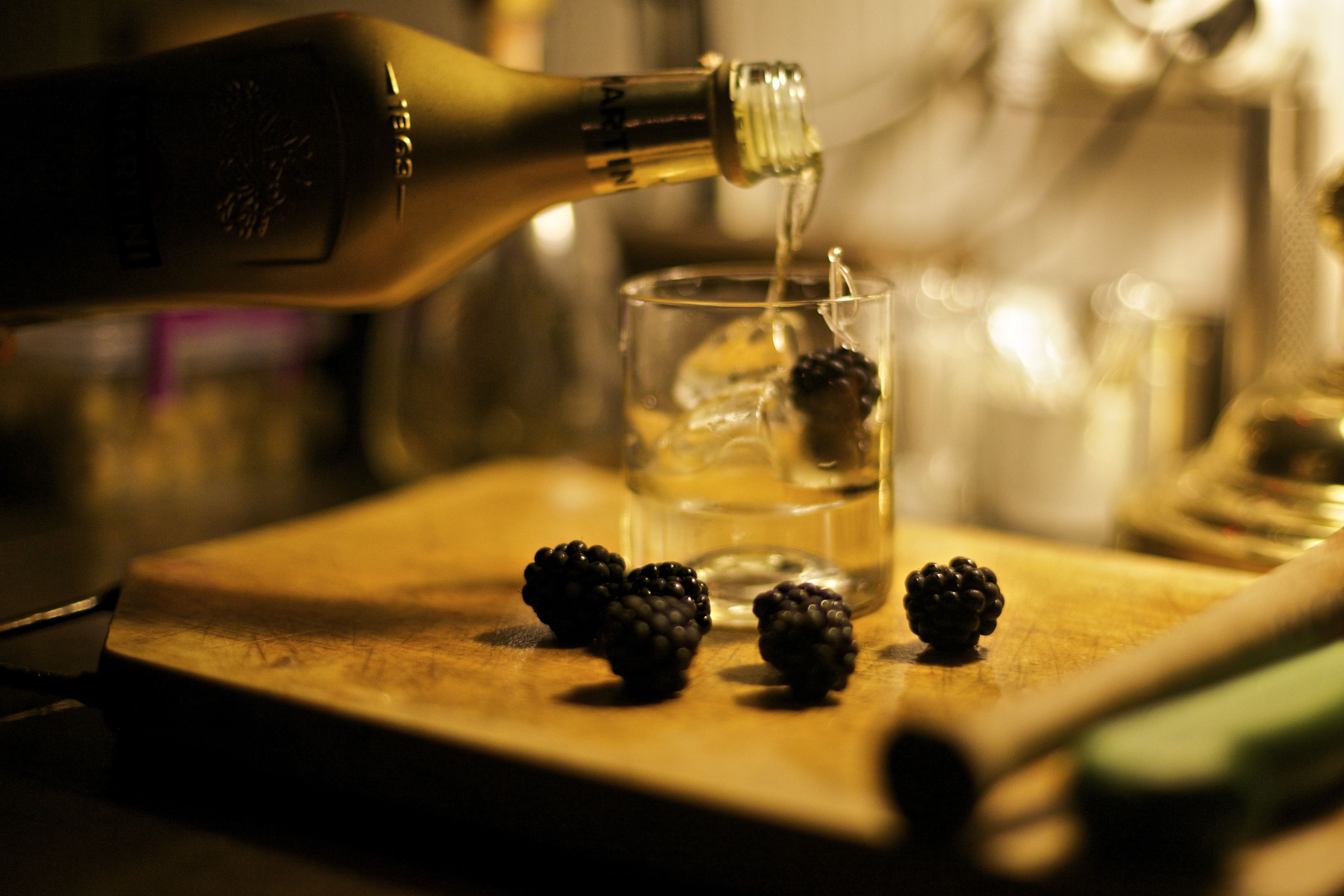 A bottle pouring liquid into a glass with blackberries on a wooden surface.