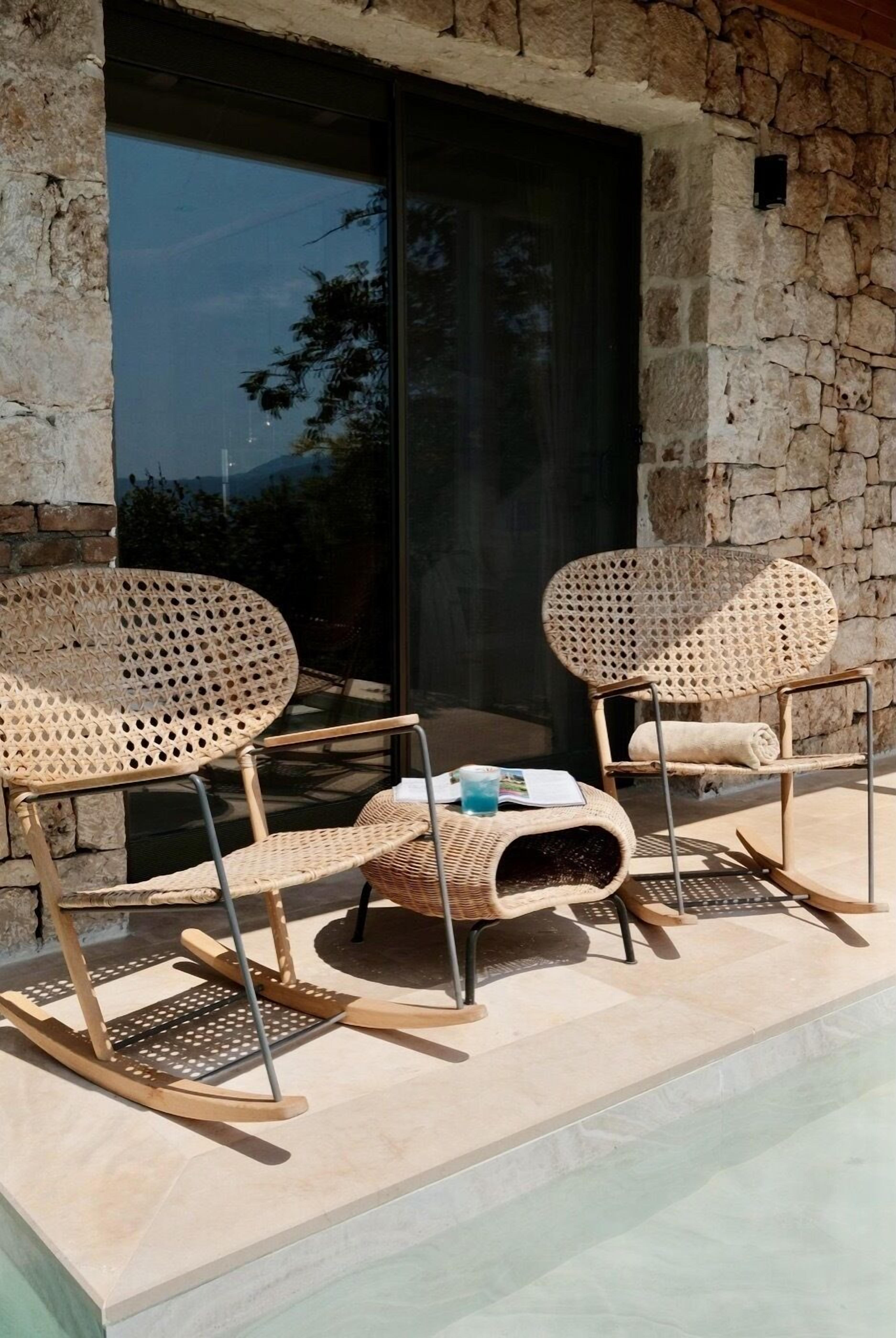 A stone wall with a glass door, wicker chairs, and a small table with a towel and a book.