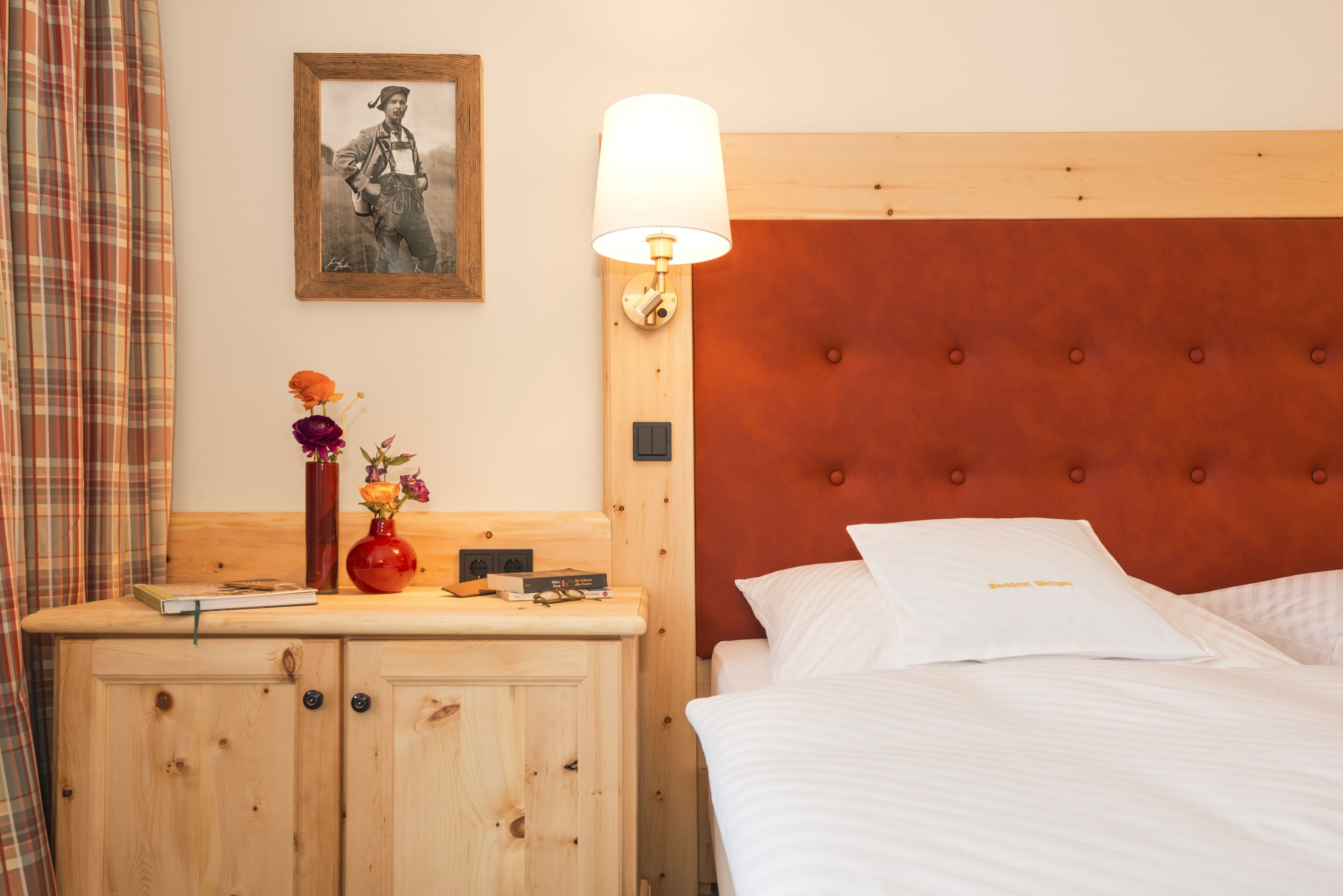 A hotel room with a wooden headboard, a bedside table with a lamp, a framed picture on the wall, and a bed with white linens.