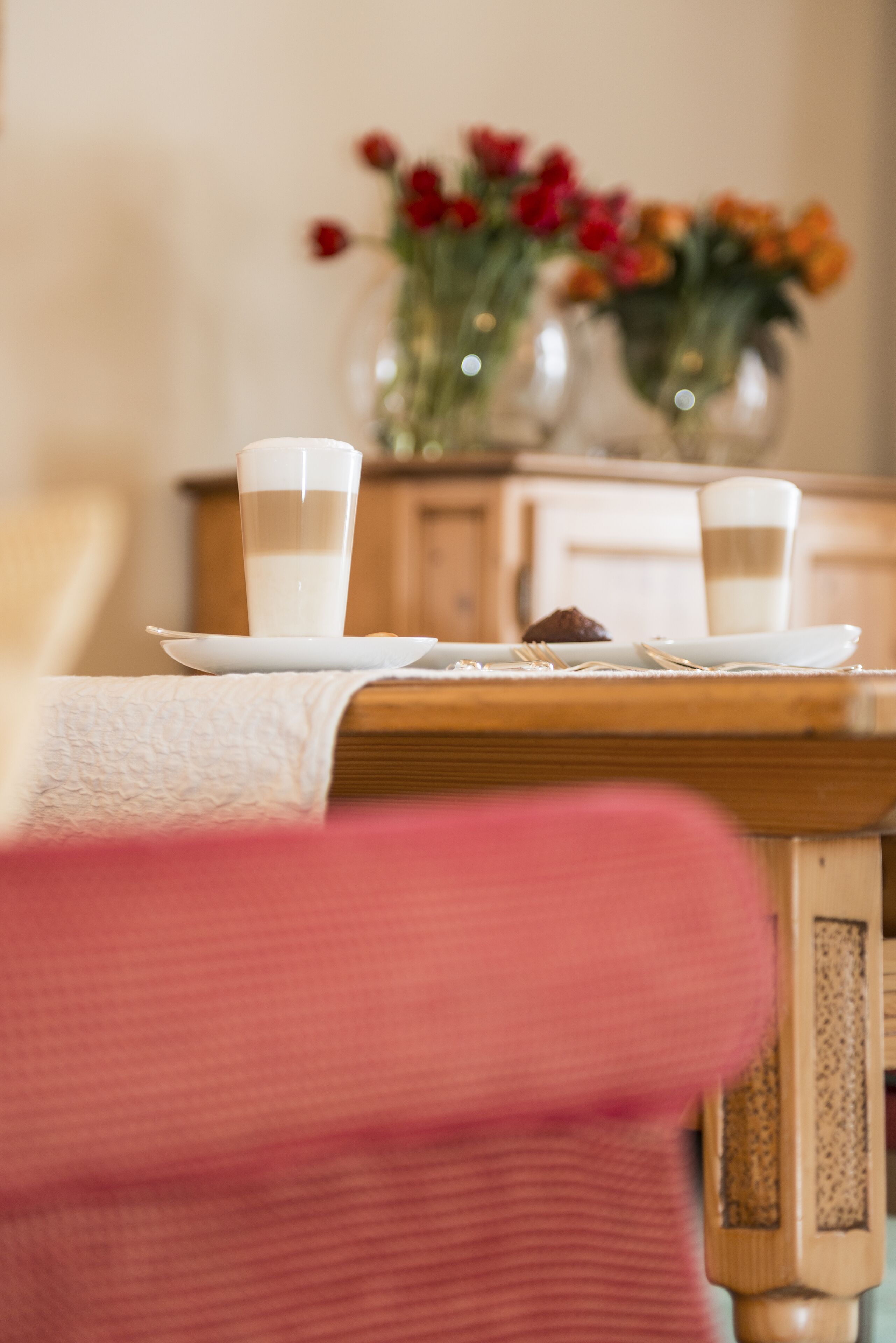 A dining table with two glasses of milk, a plate with a chocolate, and a vase with flowers in the background.