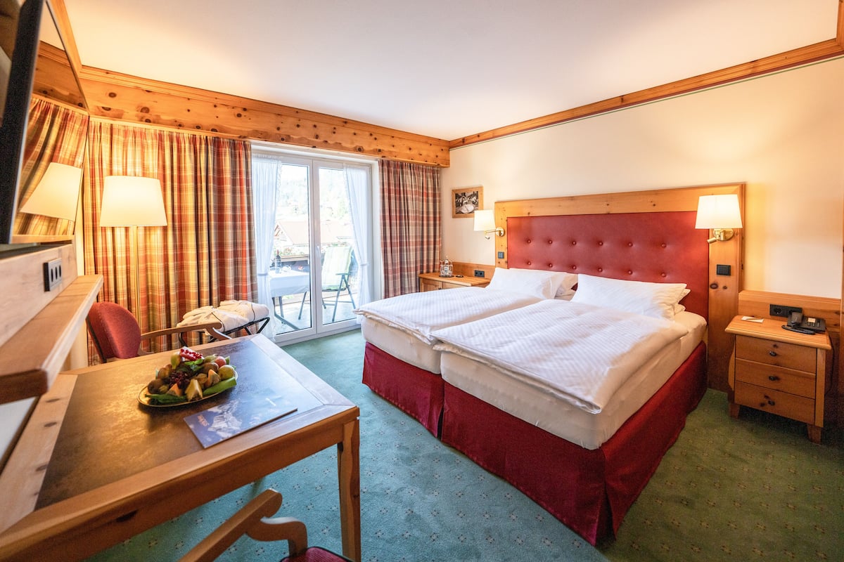 Ein Hotelzimmer mit Doppelbett, Schreibtisch mit Stuhl, einem kleinen Tisch mit Früchten und Blick ins Freie durch eine Schiebetür.