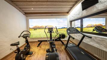 Un gimnasio en casa con una bicicleta estática, una elíptica y una cinta de correr, junto a un ventanal con vistas a la montaña.
