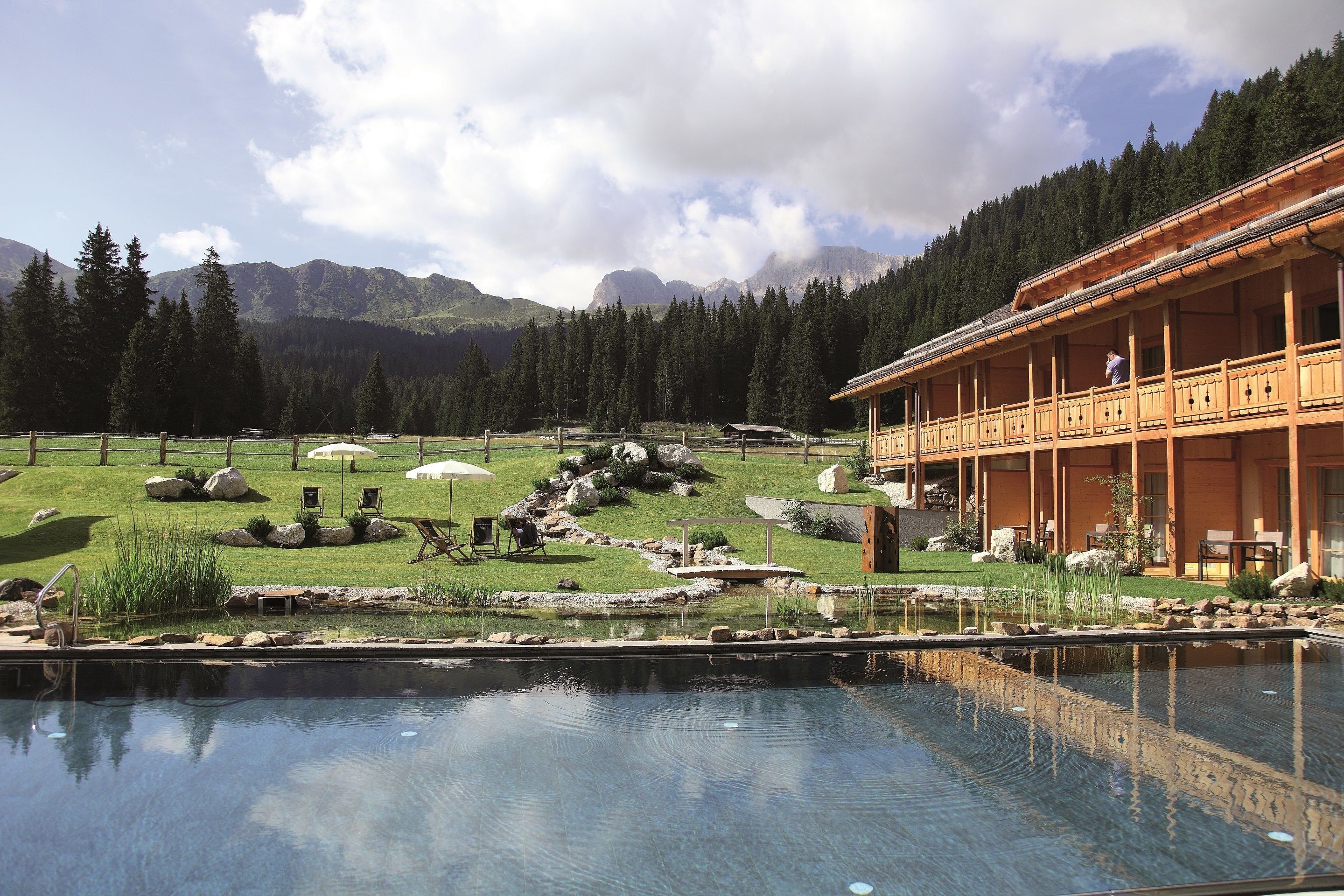 Una piscina con vistas a montañas, un edificio de madera con balcón y un césped cuidado con rocas y plantas.