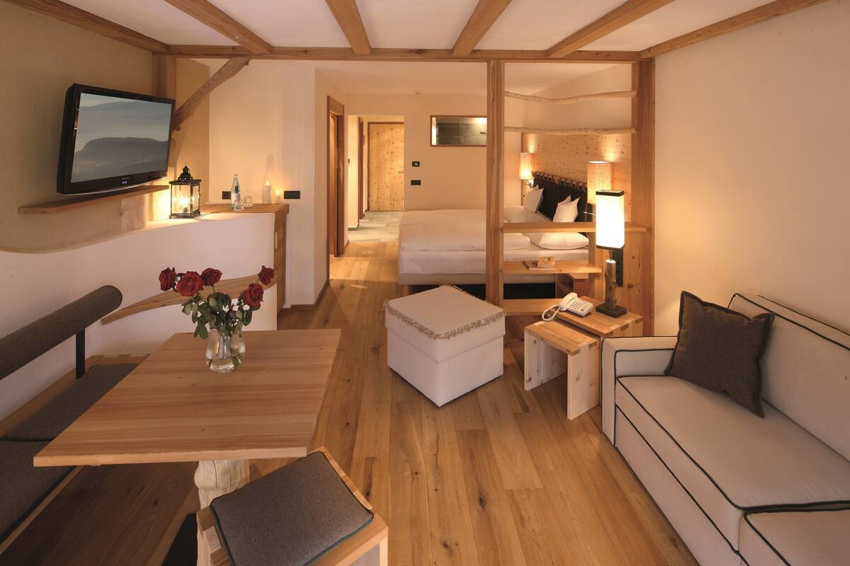 Habitación de hotel con piso de madera, una cama con cabecera, un sofá, una pequeña mesa con un jarrón de flores, un banco y un televisor montado en la pared.