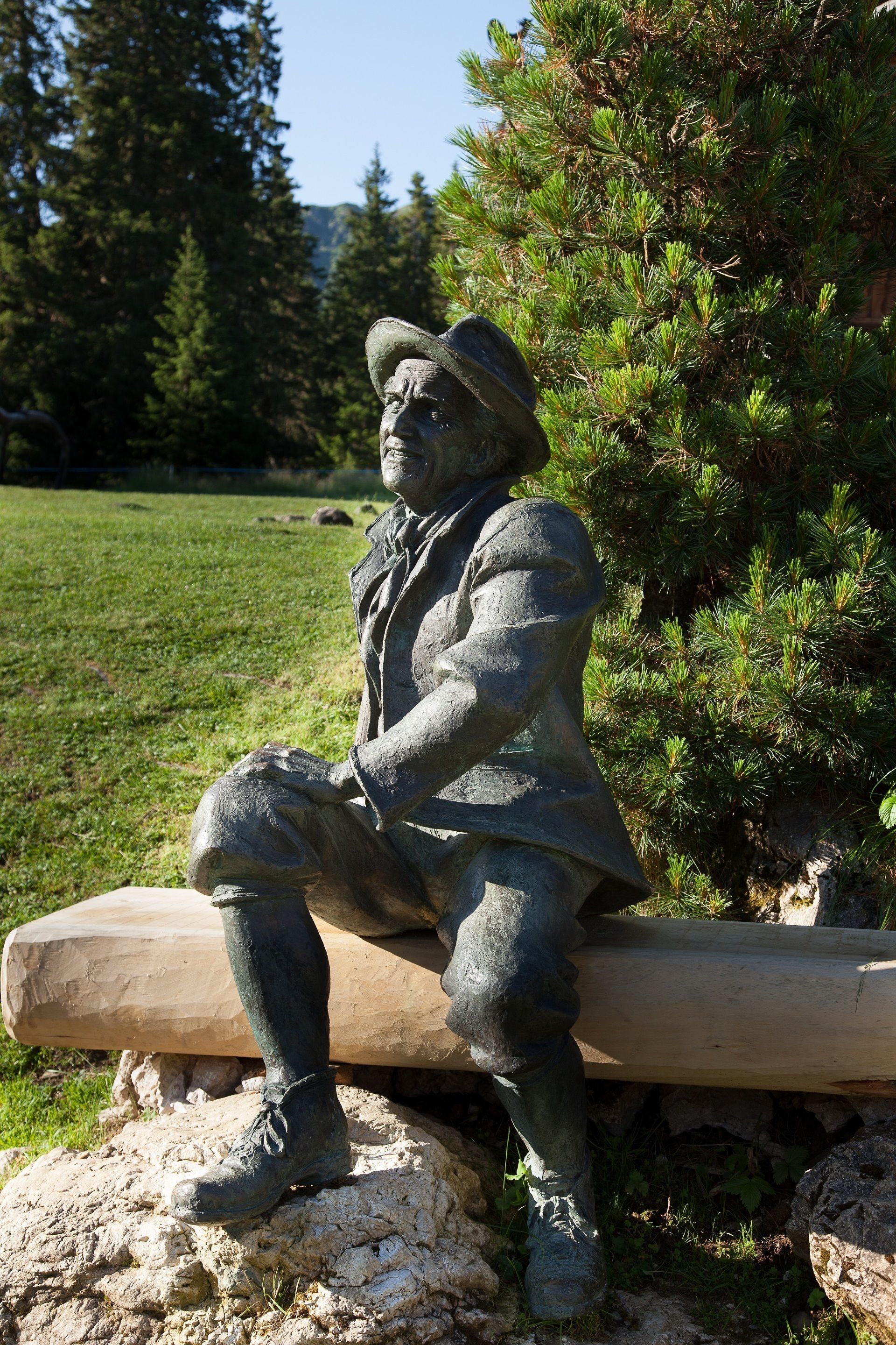 A statue of a seated man with a hat, positioned on a rock with a log bench in a park.