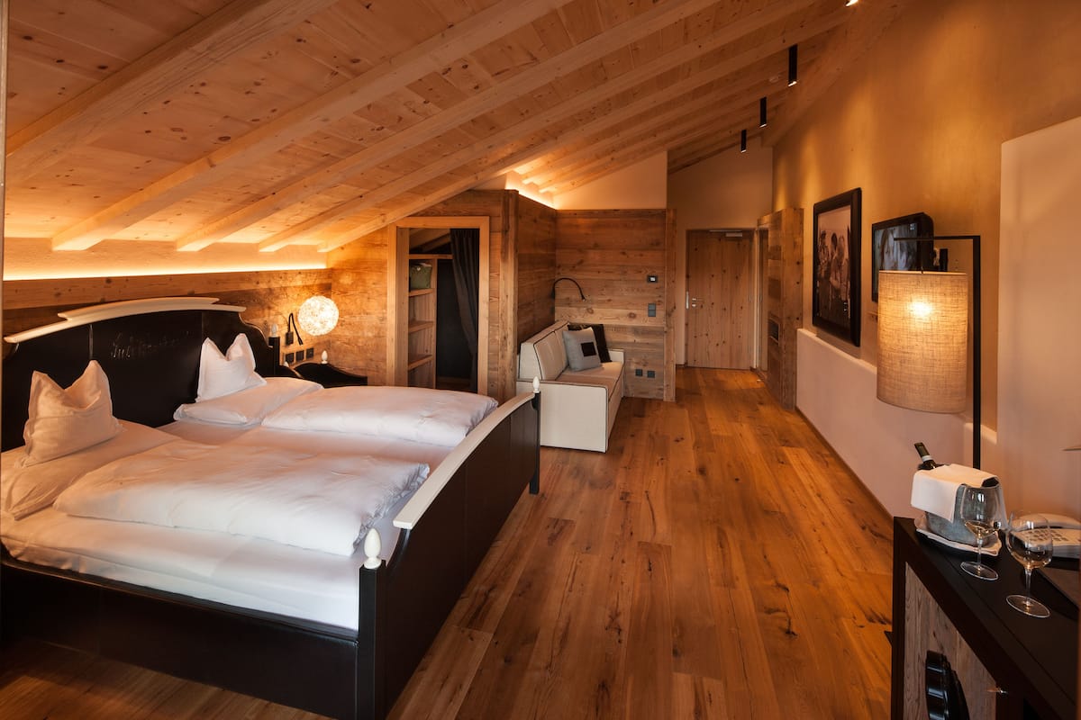 Un dormitorio con techo de madera, una cama con ropa blanca, una mesita de noche con lámpara y una pequeña zona de estar con una silla y una mesa.