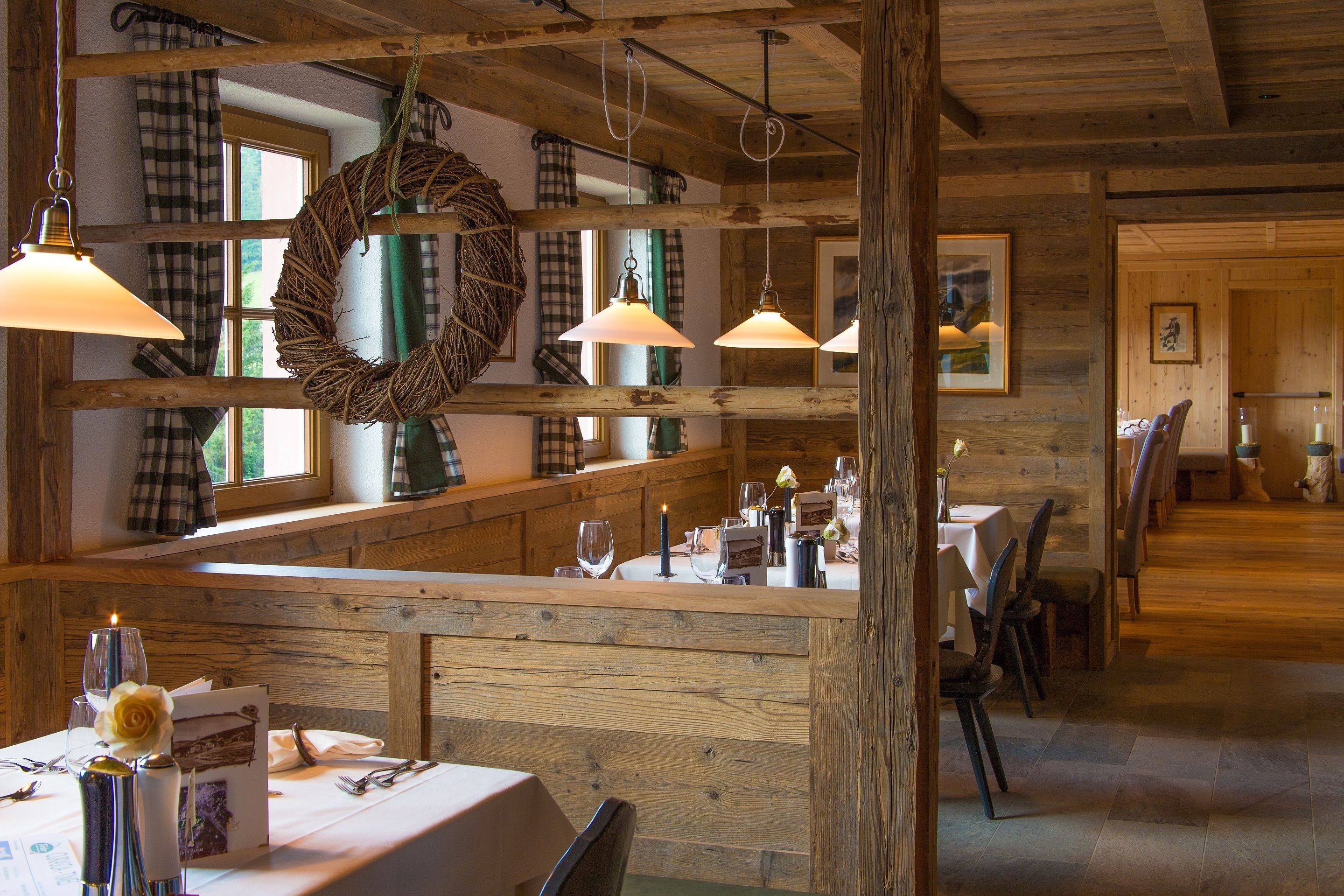 A rustic wooden interior with a long dining table set for a meal, wooden beams on the ceiling, and a large window with curtains.