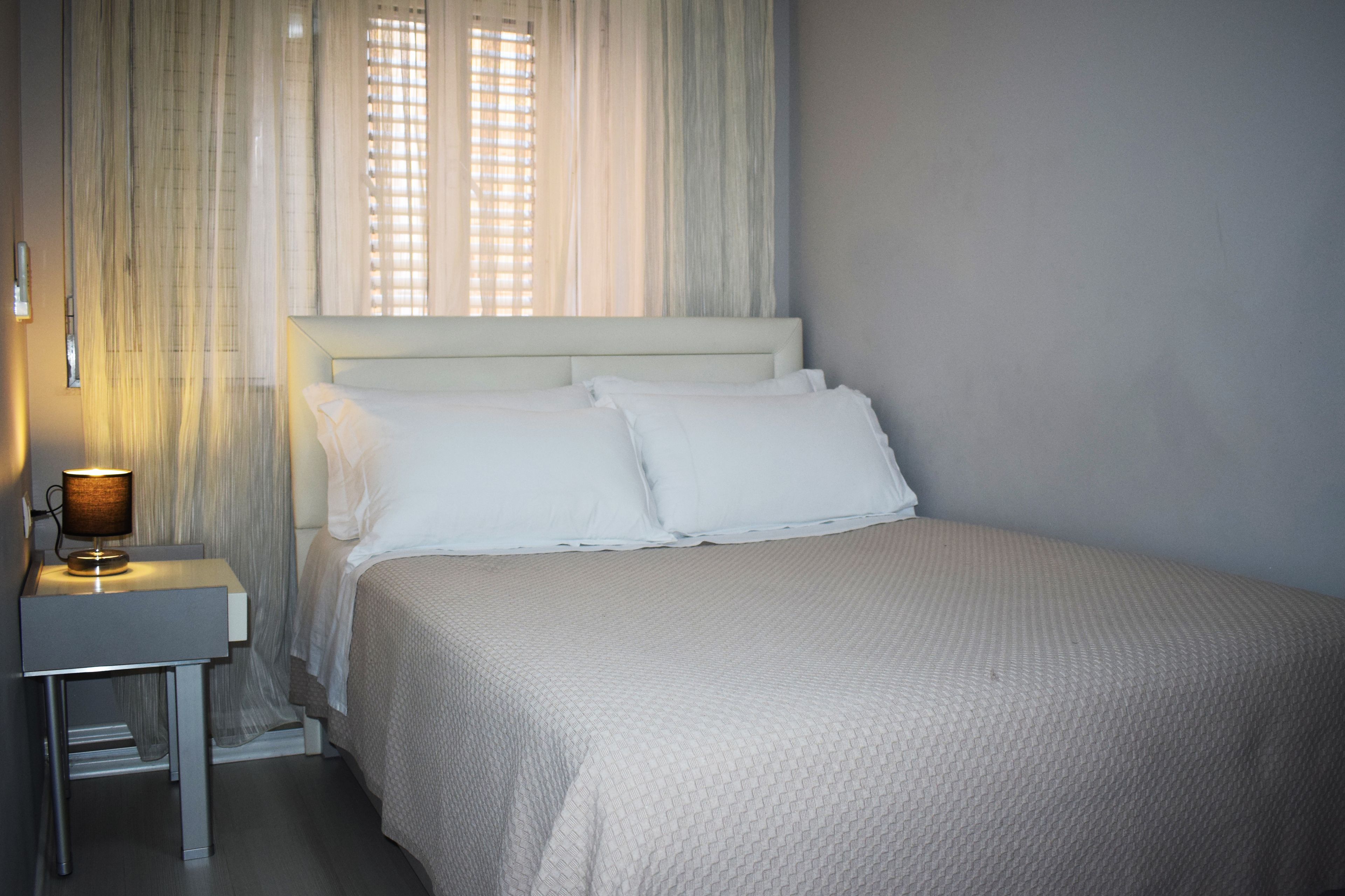 Una cama bien hecha con sábanas blancas, una mesita de noche con lámpara y cortinas traslúcidas que dejan entrar luz natural.