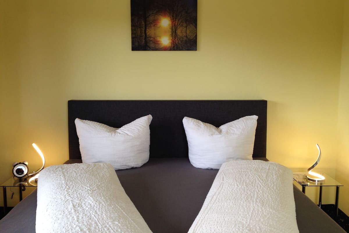 A hotel room with a large bed, two white pillows, a dark headboard, a painting of trees on the wall, and bedside lamps.