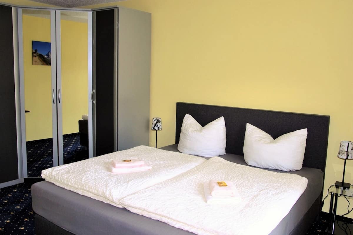 A hotel room with a bed, two pillows, a bedside table with a lamp, and a wardrobe.
