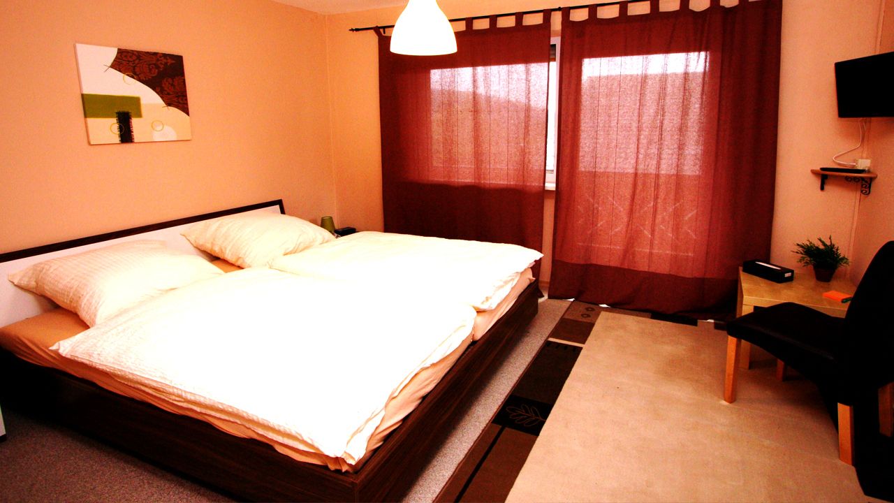 A bedroom with a bed, a desk, a chair, a TV, and a window with curtains.