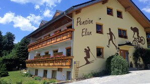 Un edificio con un balcón decorado con flores, un cartel que dice "Pension Elan" y un mural que muestra a un esquiador y un ciclista.