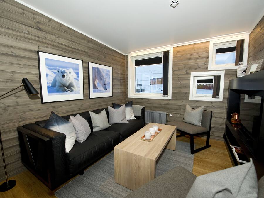 A modern living room with a black sofa, a wooden coffee table, and a TV. There are framed pictures on the wall and a floor lamp.