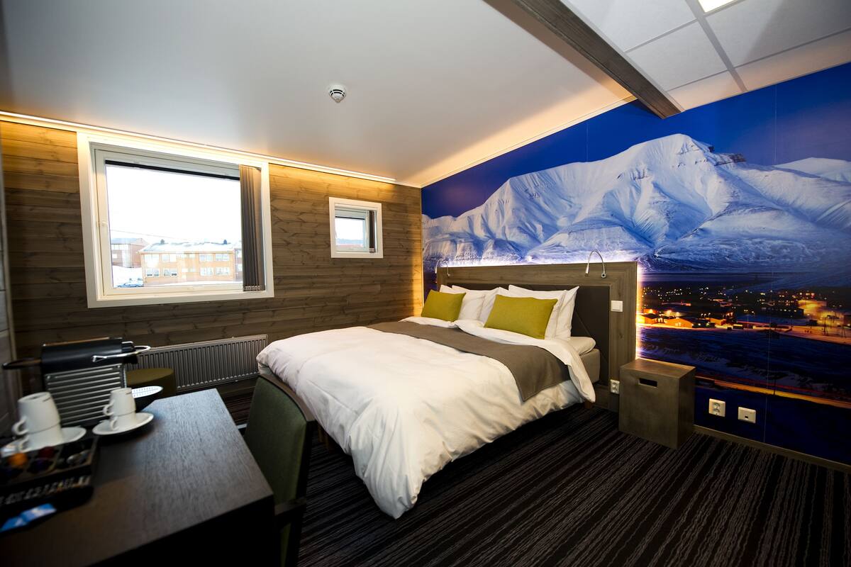 A hotel room with a bed, a desk, a chair, a window, and a wall mural of a mountain.