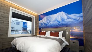 A hotel room with a large bed, a window with a view of a building, and a mural of a snowy mountain landscape.
