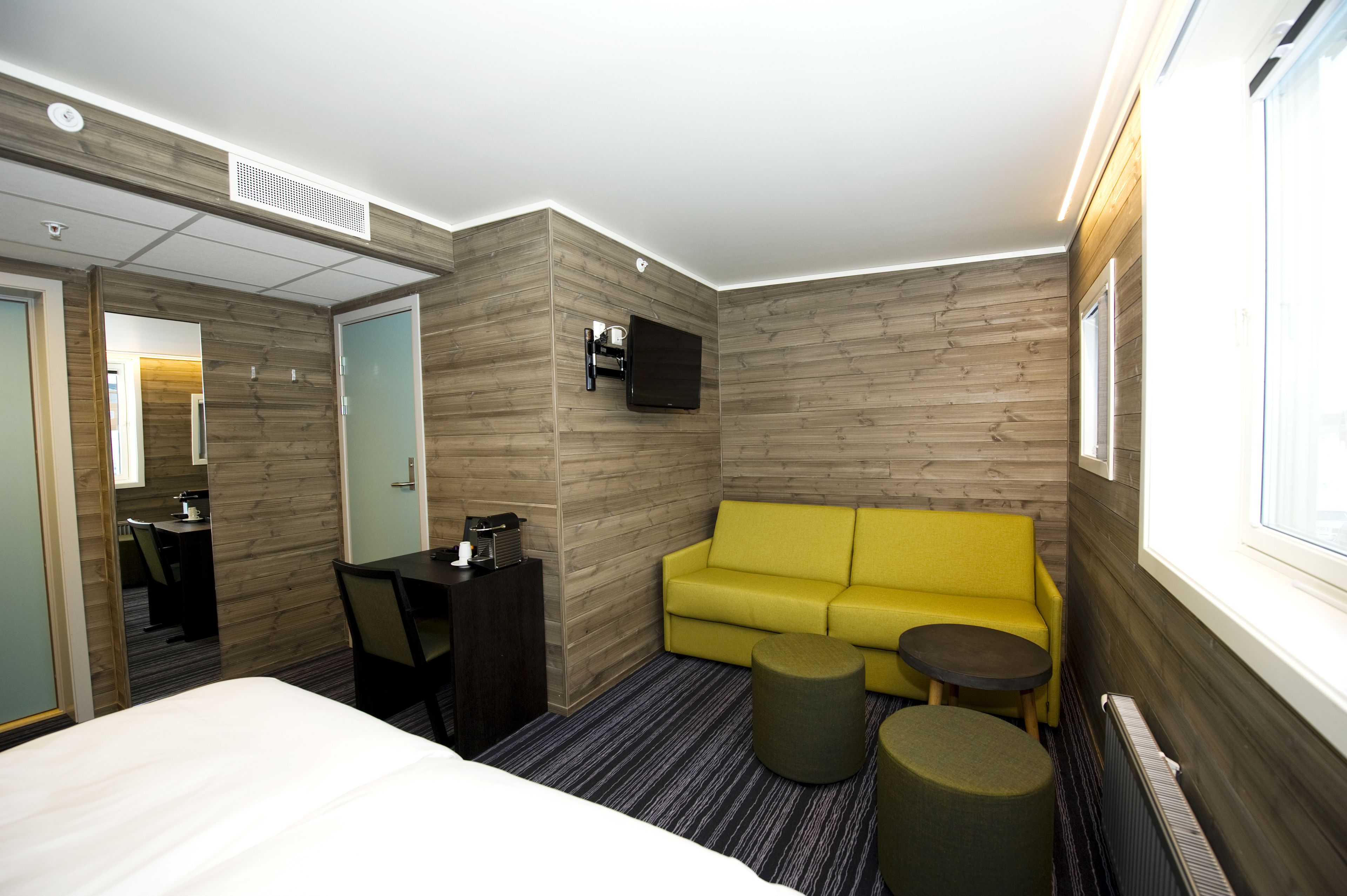Una habitación de hotel con una cama, un sofá amarillo, una mesita y un televisor montado en la pared.